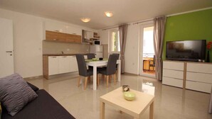 A modern living room with a sofa, a dining table with chairs, a kitchen area with cabinets and a microwave, and a TV on a stand. - Holiday apartment mit Pool (Baška)