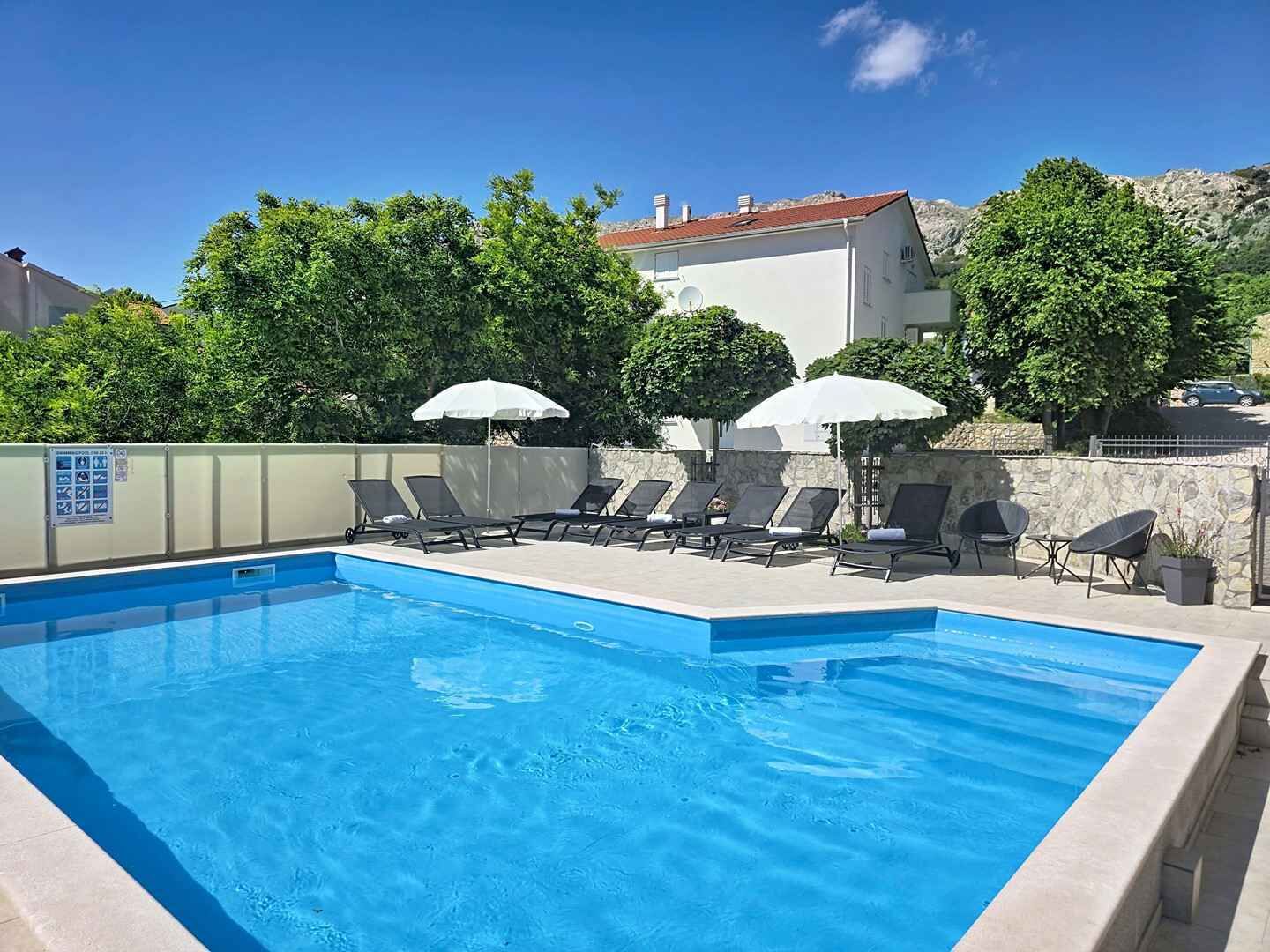Una piscina con lettini e ombrelloni, immersa nel verde e con un edificio sullo sfondo.