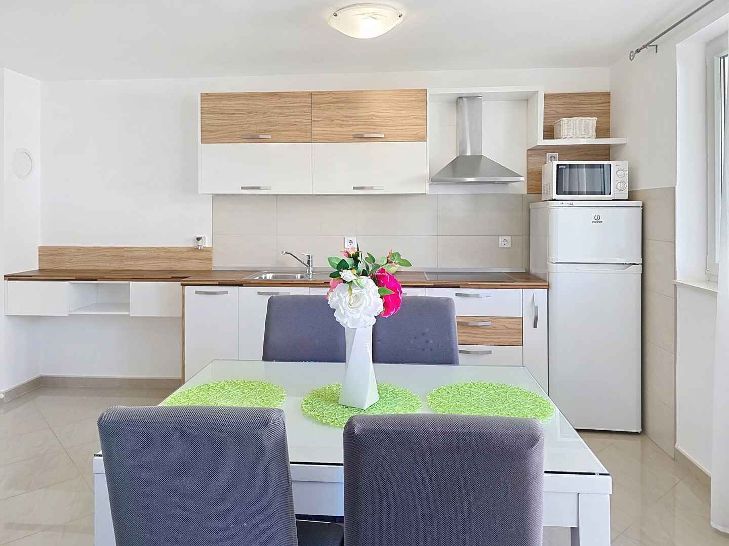 Cocina moderna con armarios blancos, extractor de acero inoxidable, microondas y refrigerador. Hay una mesa de comedor con dos sillas y un florero con flores.