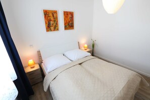 Ein ordentlich gemachtes Bett mit einer beigen Decke, zwei weißen Kissen und einem hölzernen Kopfteil. Auf jeder Seite des Bettes stehen je eine Nachttischlampe, darüber an der Wand hängen zwei abstrakte Gemälde.