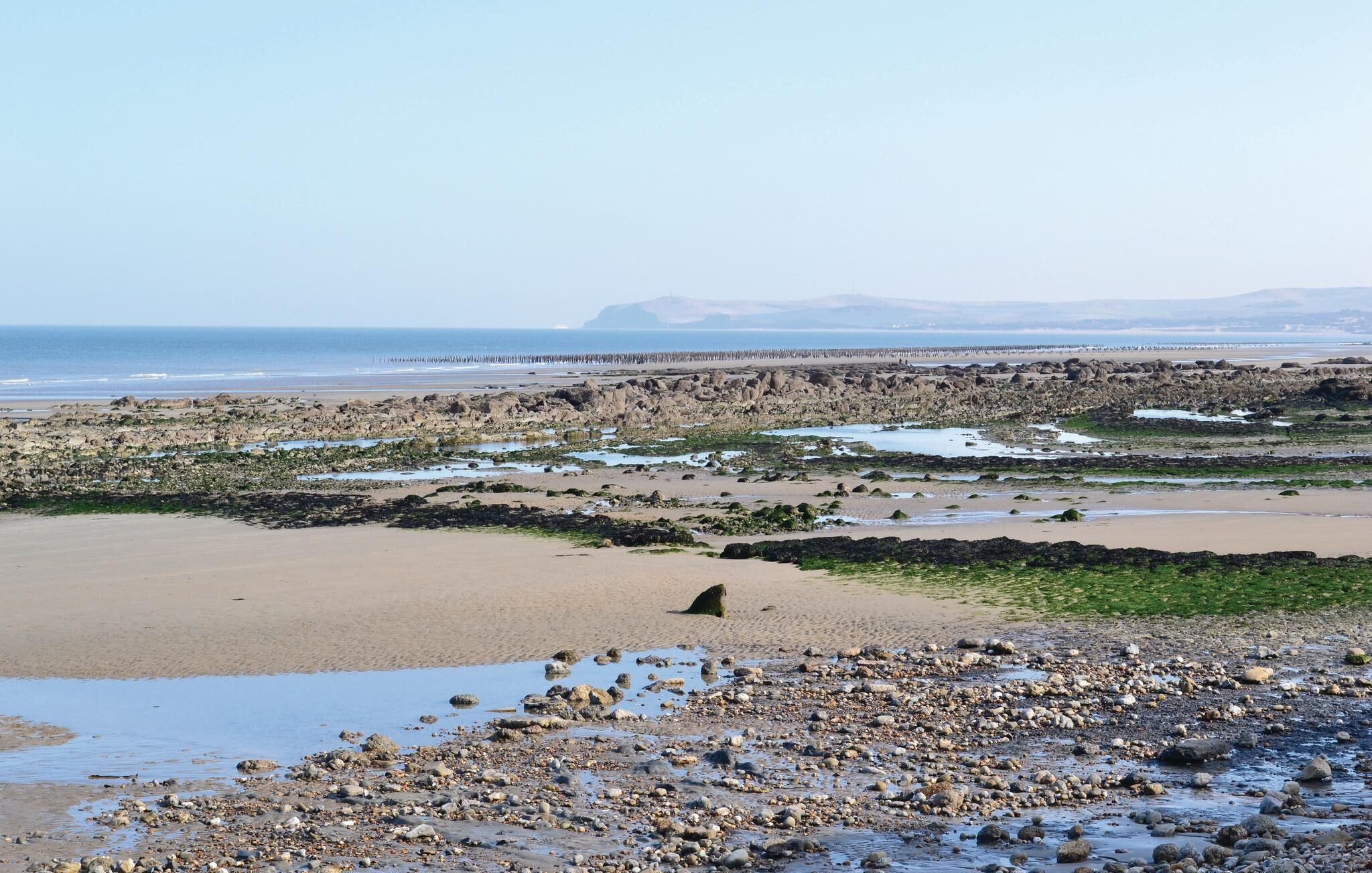 Une plage rocheuse avec une zone de sable et une jetée visible au loin.
