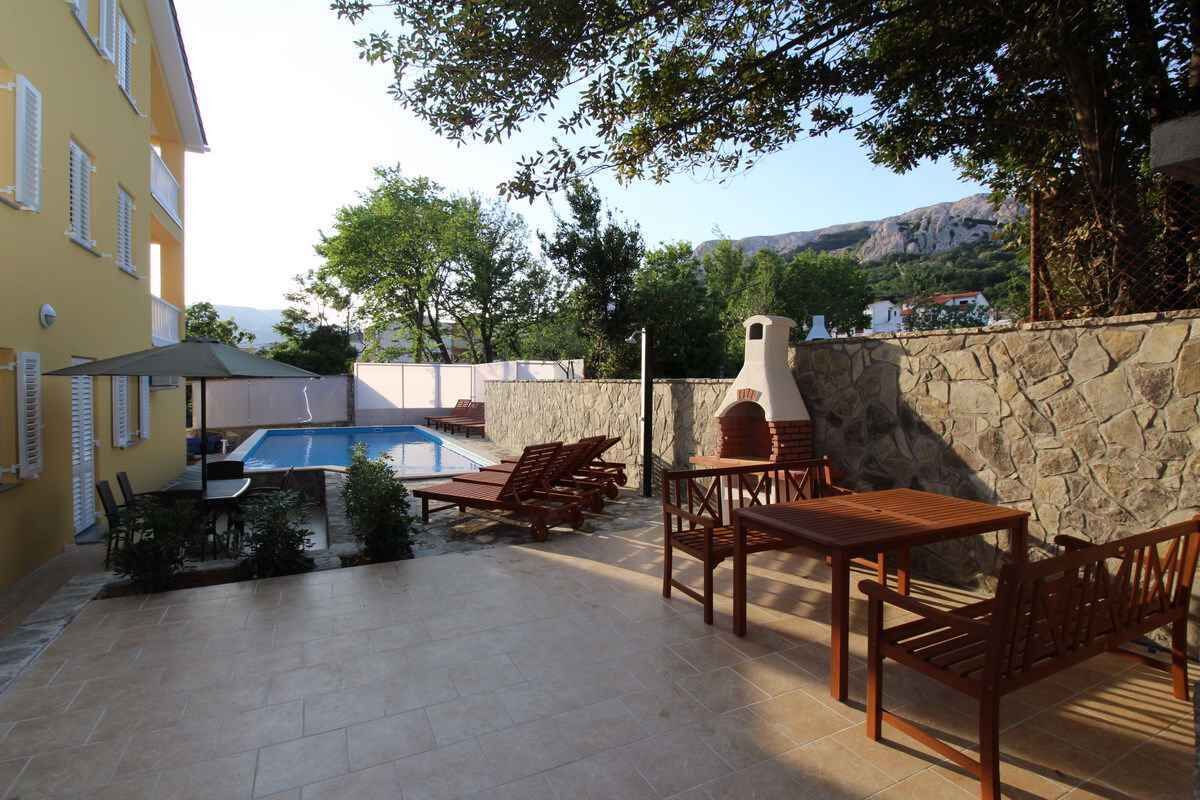 A poolside area with a stone wall, wooden furniture, and a view of the mountains.