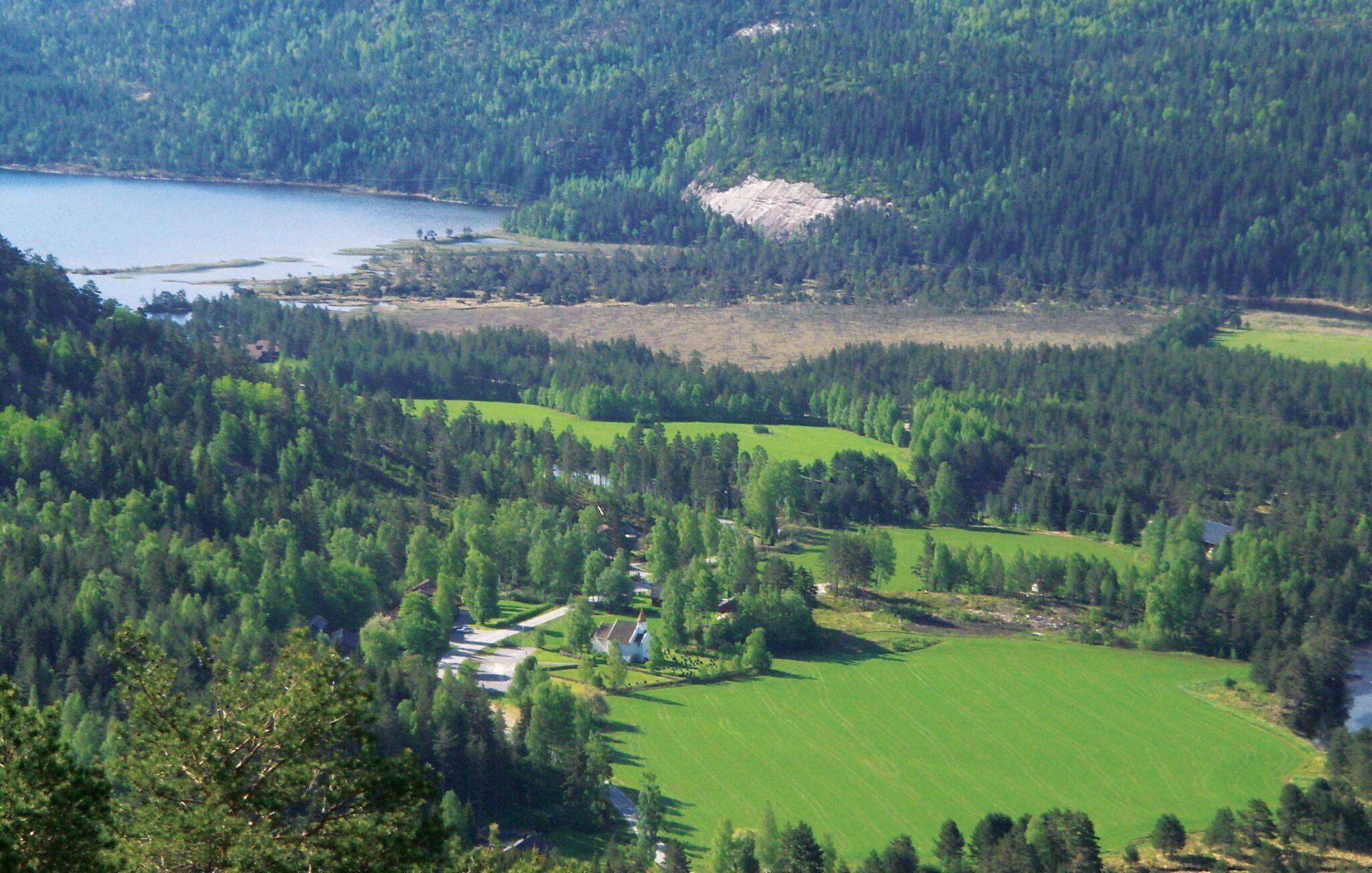 Paisatge rural amb un llac, un bosc dens i un petit grup d'edificis.