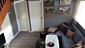 A cozy living room with a grey sofa, a small table, and a black ottoman. There are framed pictures on the wall and a wooden staircase in the background.