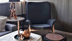 A modern living room with a dark blue armchair, a round wicker table with a metal tray, a white vase, and a textured ottoman.