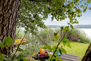Et picnicbord med en frugtkurv og juiceflasker foran en sø og træer.