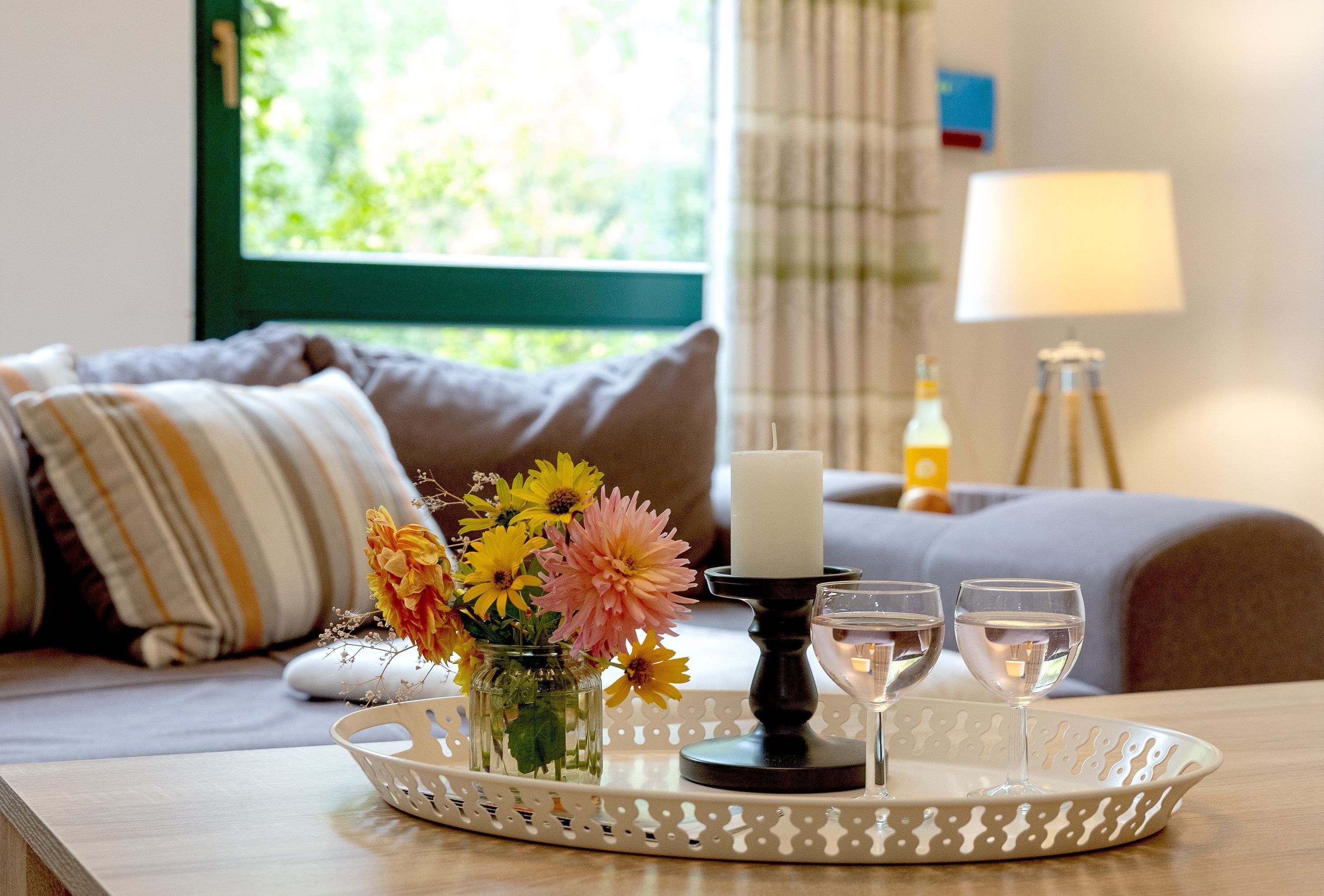 A tray with a candle, two glasses of water, and a vase of flowers on a table in a room with a bed and a lamp in the background.