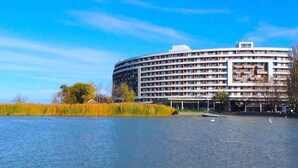 Un grand hôtel à plusieurs étages, de forme circulaire, situé près d’une étendue d’eau, avec des bateaux et des roseaux au premier plan.