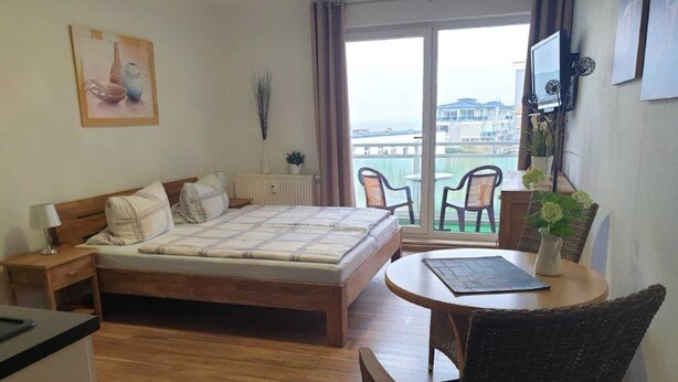 A hotel room with a bed, a desk, a chair, a small table, and a balcony with a view. - (157) 1- room apartment - apartment complex Yachthafenresidenz (Kühlungsborn)