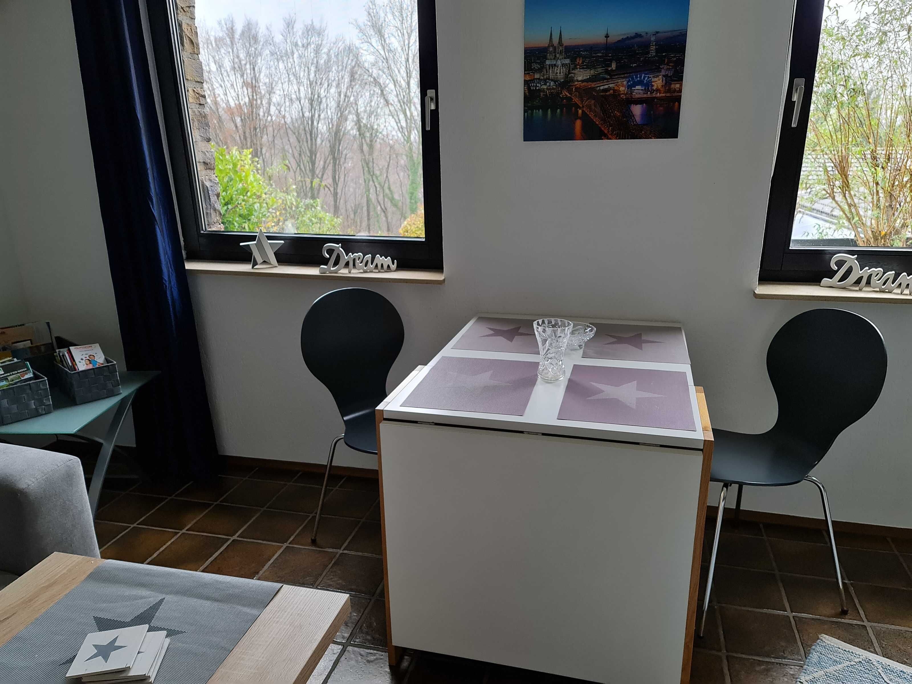 Ein Zimmer mit Tisch, zwei Stühlen, einem Fenster mit Blick auf Bäume und eine Stadtansicht sowie einer kleinen Ablagefläche mit einem dekorativen Gegenstand.
