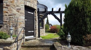 Un edificio de piedra con puerta negra, una pérgola de madera y un jardín bien cuidado.
