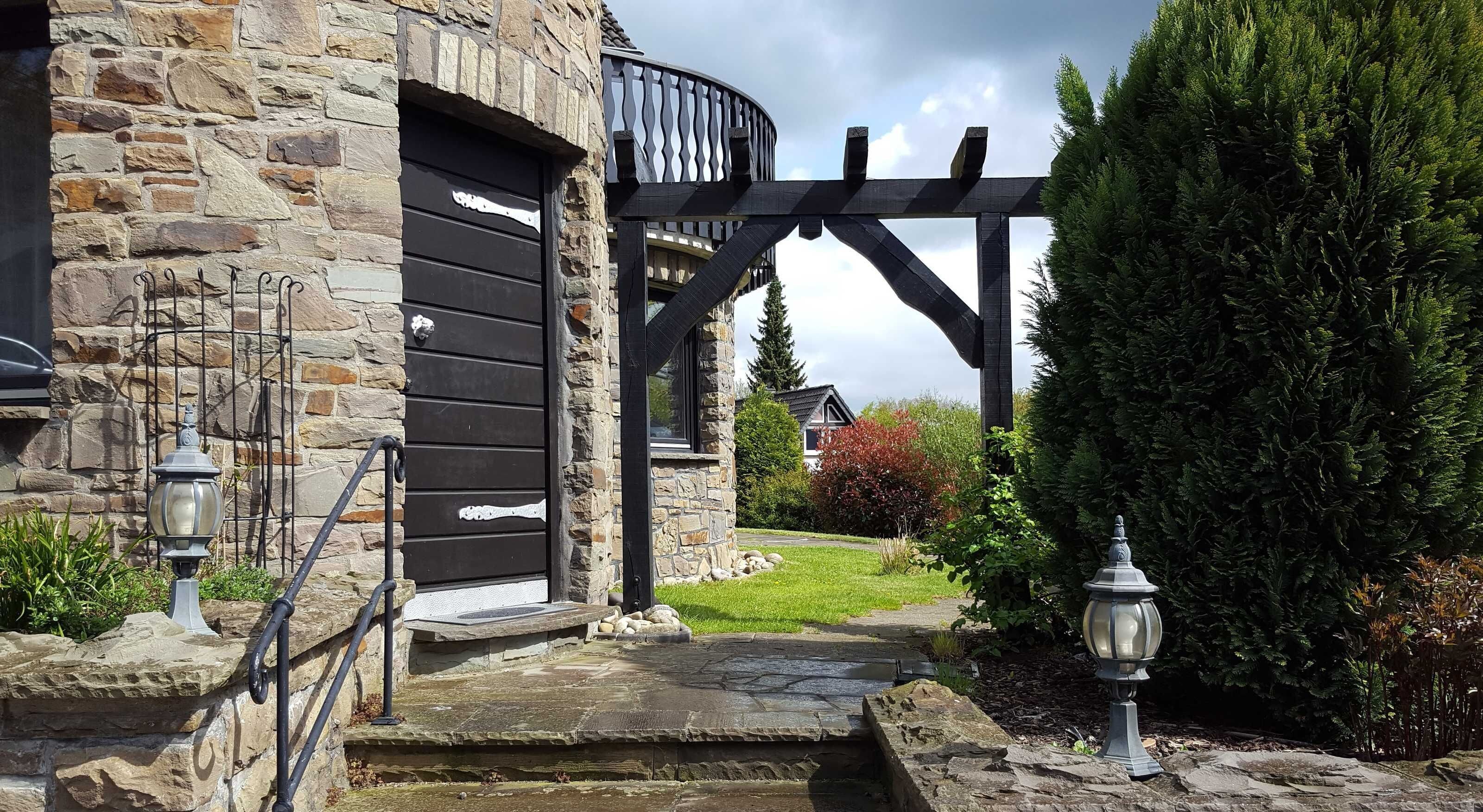 Ein Steinhaus mit einer schwarzen Tür, einer Holzpergola und einem gepflegten Garten.