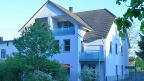 A two-story house with a balcony on the upper floor, surrounded by greenery and a metal fence. - Apartment 3 - Bluehouse - Apartments Egonschultz (Zempin)
