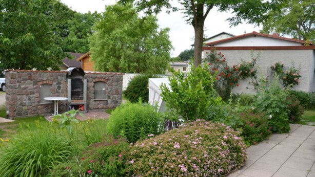 A stone barbecue area with a table and chairs, surrounded by a garden with various plants and flowers. - Apartment 3 - Bluehouse - Apartments Egonschultz (Zempin)