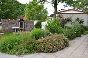 A stone barbecue area with a table and chairs, surrounded by a garden with various plants and flowers.