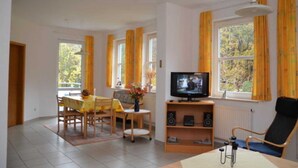 A living room with a television, a dining table with chairs, a sofa, and a window with curtains. - Apartment 3 - Bluehouse - Apartments Egonschultz (Zempin)