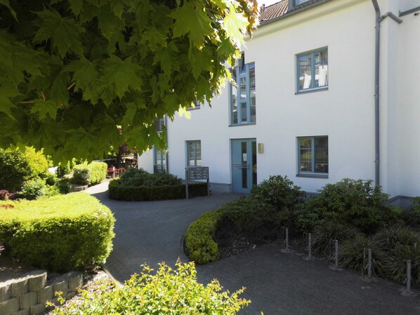 A white building with blue-framed windows, a gated entrance, and a well-maintained garden with hedges and trees.