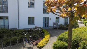 Un edificio con entrada pavimentada, una bicicleta estacionada afuera y un jardín bien cuidado con diversas plantas.