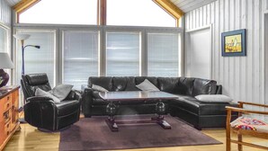 Ein gemütliches Wohnzimmer mit Ledersofa, Couchtisch aus Holz und Holzstuhl. Große Fenster mit Jalousien, eine Lampe und ein gerahmtes Bild an der Wand.
