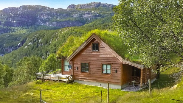 Eine Holzhütte mit geneigtem Dach, umgeben von üppiger Vegetation und Bergen im Hintergrund.