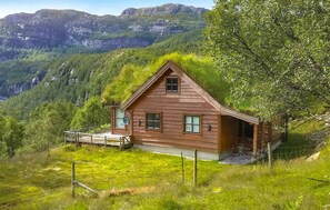 En stuga byggd av trä med ett sluttande tak, omgiven av frodig grönska och med berg i bakgrunden.