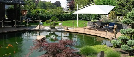 Um lago com carpas, uma plataforma de madeira e área de estar, rodeado por vegetação e um gazebo.