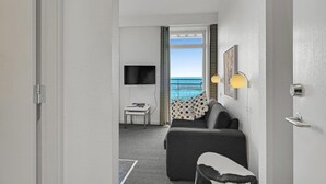 A hotel room with a grey sofa, a small table, a TV, and a view of the sea through a window.