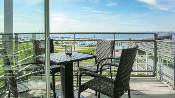 A balcony with wicker chairs and a table, overlooking a marina with boats and a distant shoreline.