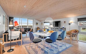 A modern living room with a wooden ceiling, a dining area with blue chairs, and a large window offering a view of the outdoors. - 3 bedroom beautiful home in Hjørring (Hjørring)