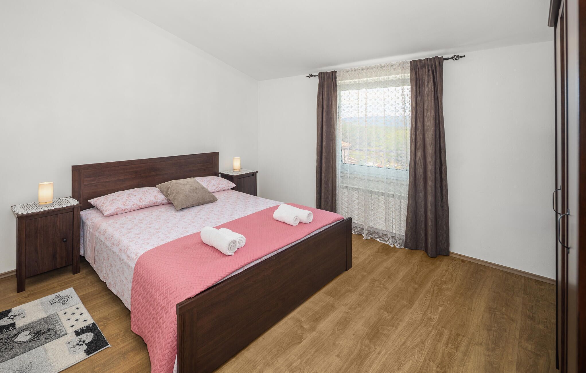 Un dormitorio ordenado con una cama de madera, una mesita de noche con lámpara, una ventana con cortinas y una alfombra estampada en el suelo.