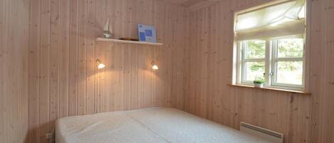 A room with wooden walls, a bed with a white bedspread, a window with a view of greenery, and two wall-mounted lights.