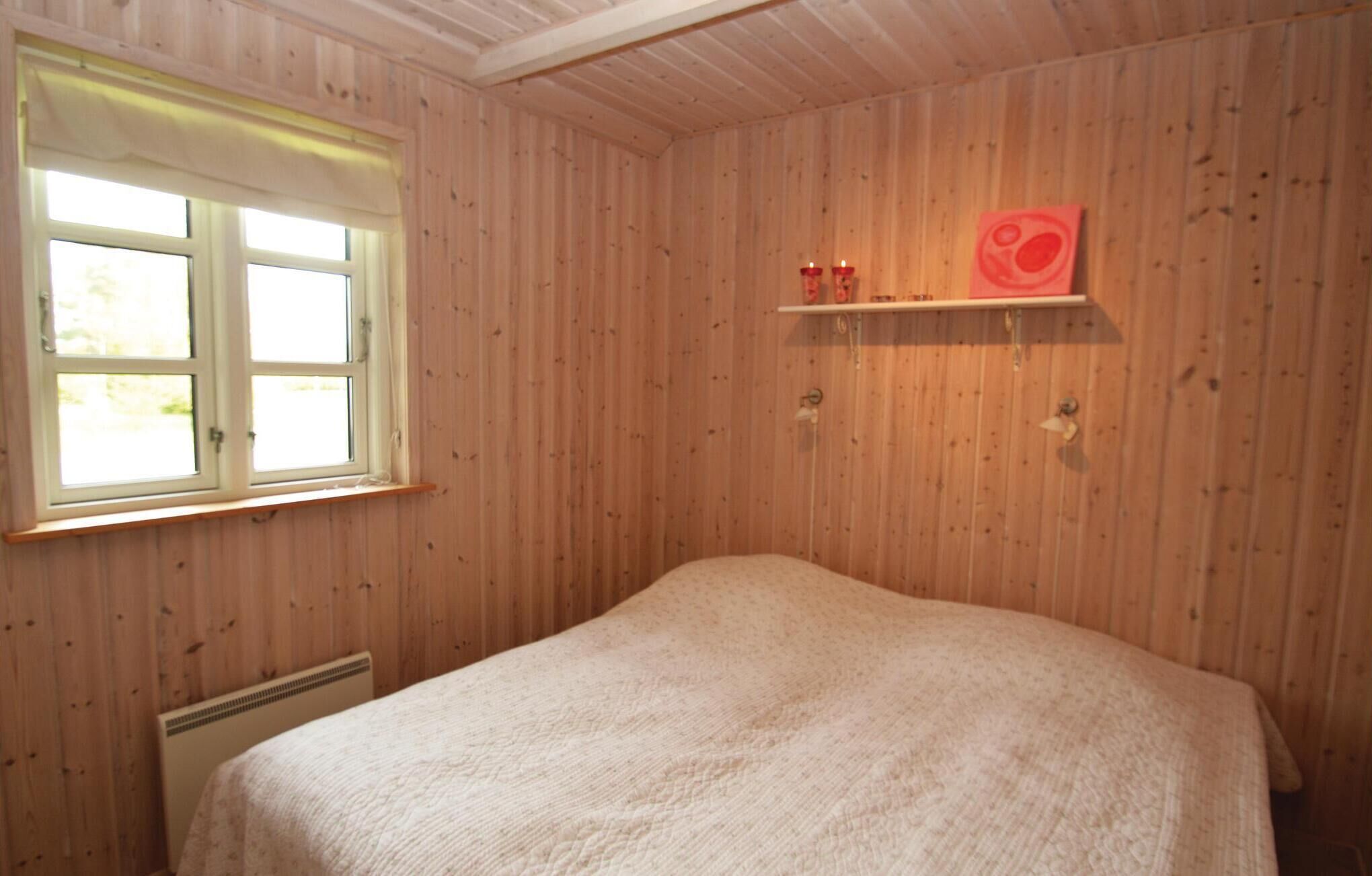 A room with wooden paneling, a bed with a white blanket, a window with white shutters, and a shelf with two red candles.