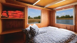 Une cabane comprenant un lit, une étagère avec des objets rouges et deux fenêtres donnant sur un lac et des arbres.