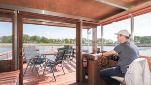 Un pont de bateau avec une table dressée pour le repas, une roue de barre et une vue sur une rivière paisible.