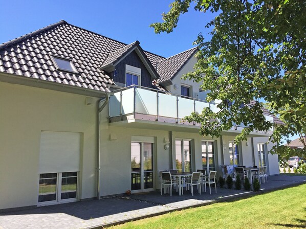 A two-story residential building with a balcony, large windows, and a patio area with outdoor furniture.