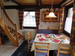 A dining area with a table set for four, wooden chairs, a radiator, a window with curtains, and a staircase.