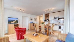 A modern living room with a wooden coffee table, red sofa, and blue sofa. There is a dining area with wicker chairs and a kitchen with white cabinets and appliances. - Villa Aquamarina Whg. 13, BALCONY w. LAKE VIEW, CHAIR - Villa Aquamarina, 1st floor (Heringsdorf)