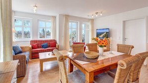 A cozy living room with a wooden table, wicker chairs, a red plaid sofa, and a television displaying a beach scene. - Villa Aquamarina Whg. 13, BALCONY w. LAKE VIEW, CHAIR - Villa Aquamarina, 1st floor (Heringsdorf)