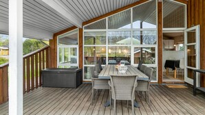 Et overdækket udendørs spiseområde med bord og stole, store vinduer samt udsigt til baghaven.