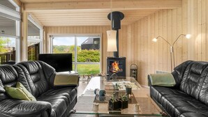 En hyggelig stue med to lædersofaer, et glasbord, en pejs og et stort vindue med udsigt over grønne omgivelser.