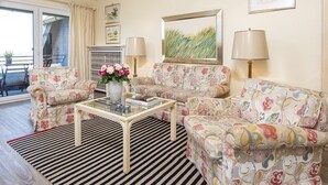 A living room with floral-patterned sofas, a glass coffee table, and two lamps. There is a painting on the wall and a window with curtains. - Nautilus 12 from A&M - Nautilus 12 (Kampen (Sylt))