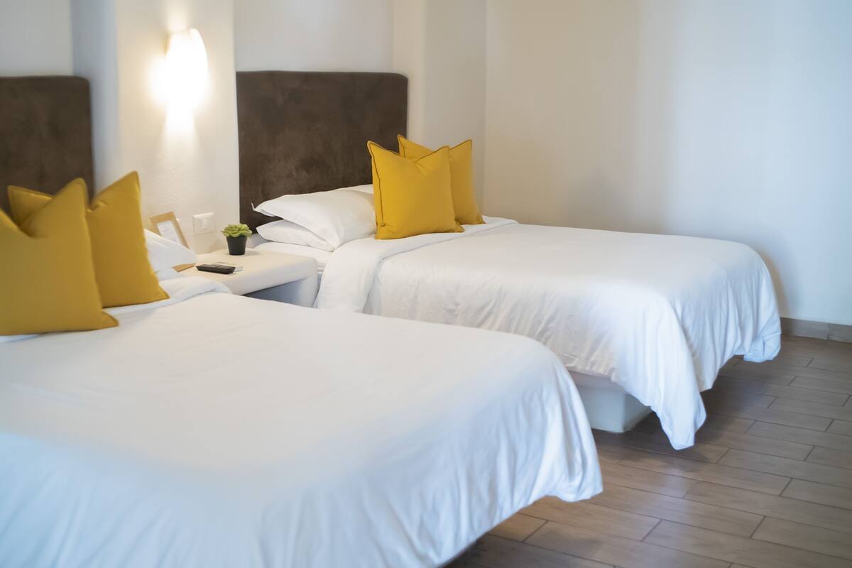 Hotellihuone, jossa on kaksi sänkyä, ja molemmissa on valkoiset vuodevaatteet sekä yksi keltainen tyyny.