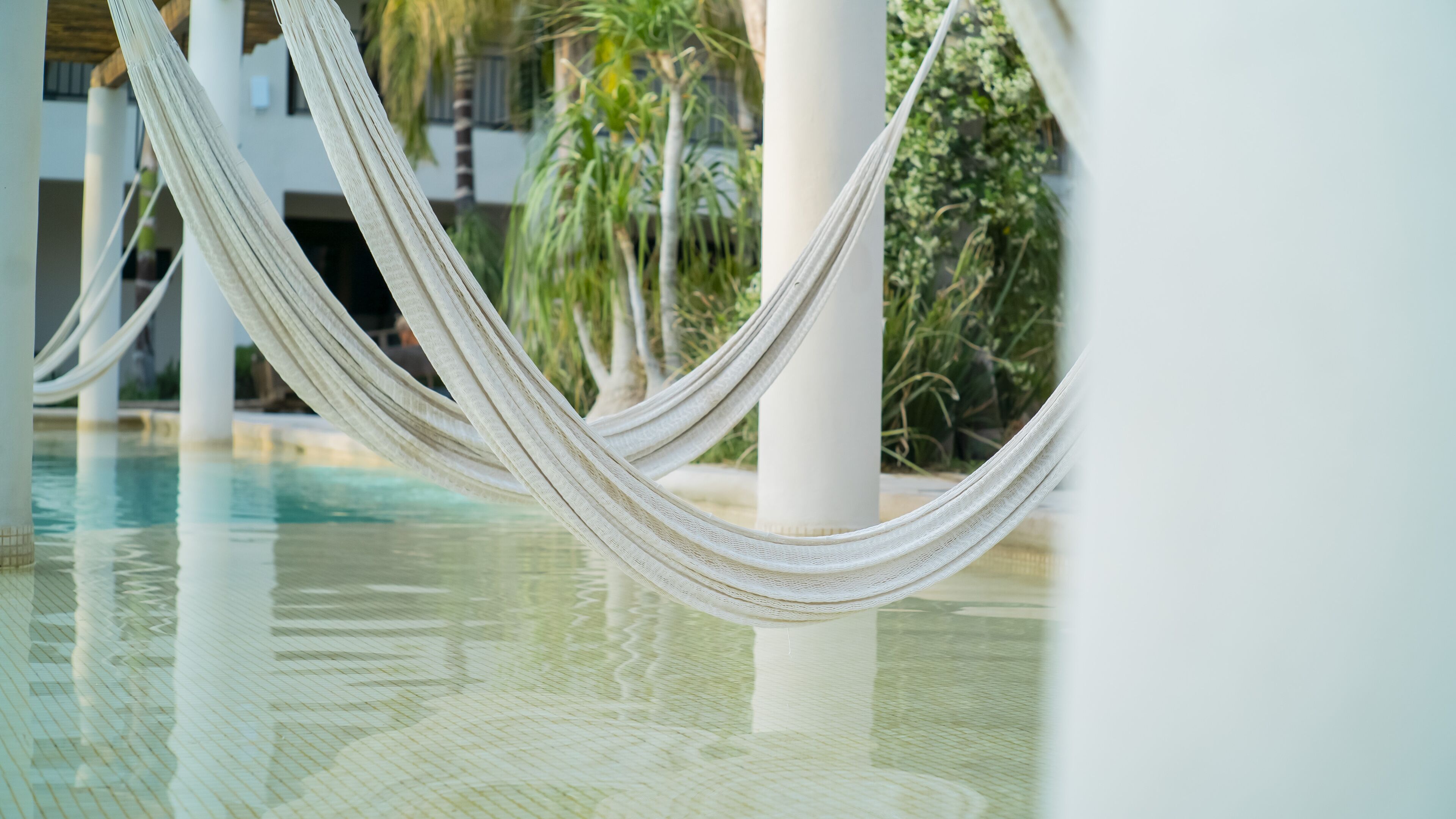 Eine Hängematte über einem Pool mit Blick auf Palmen und ein Gebäude im Hintergrund.