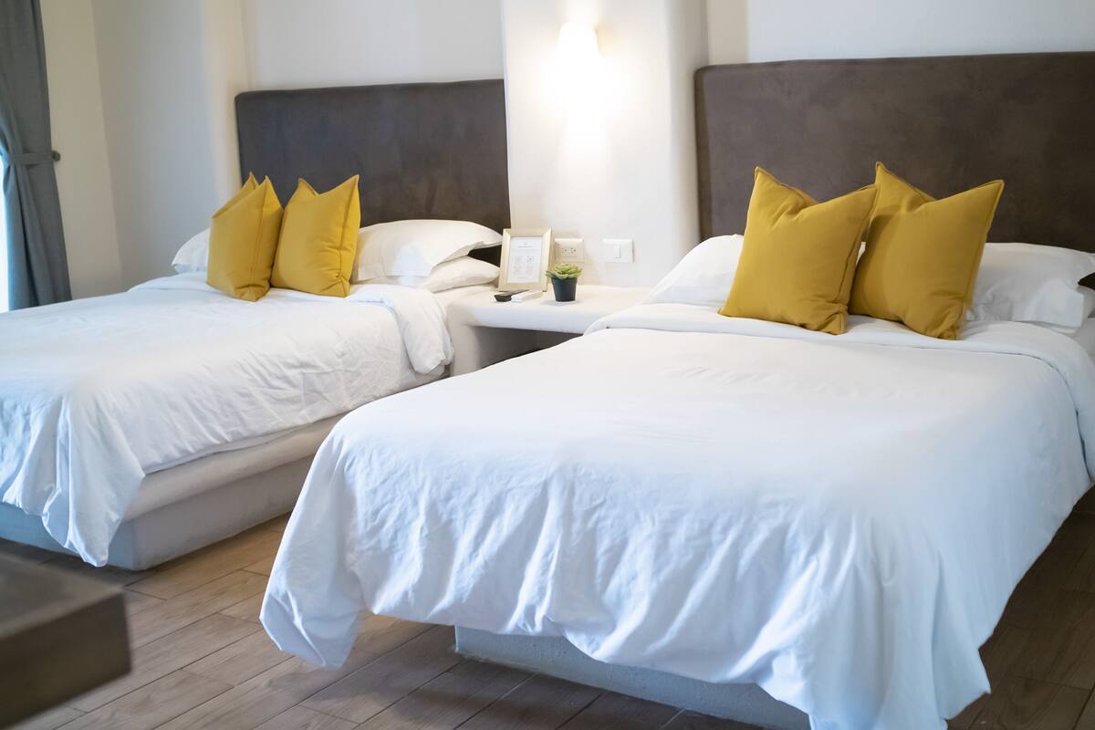Hotellihuoneessa on kaksi sänkyä, joissa on valkoiset lakanat ja keltaiset tyynyt. Huoneessa on yöpöytä, jossa on lamppu ja pieni kasvi.