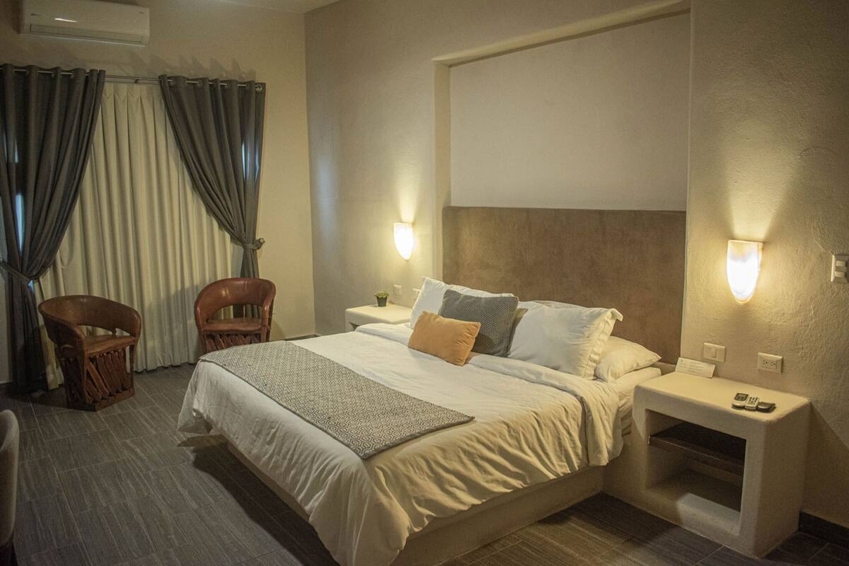 Hotellihuone, jossa on suuri sänky, kaksi tuolia, yöpöytä lampun kanssa ja pieni sivupöytä, jossa on puhelin.