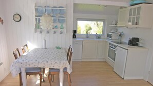 Een eetgedeelte met een tafel voor vier personen, een keuken met witte kasten en apparatuur, en een klok aan de muur.