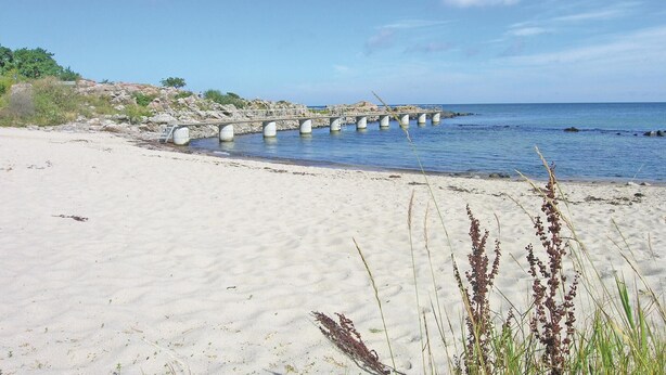 Een strand met een betonnen steiger die het water in reikt, omringd door zand en schaarse begroeiing.