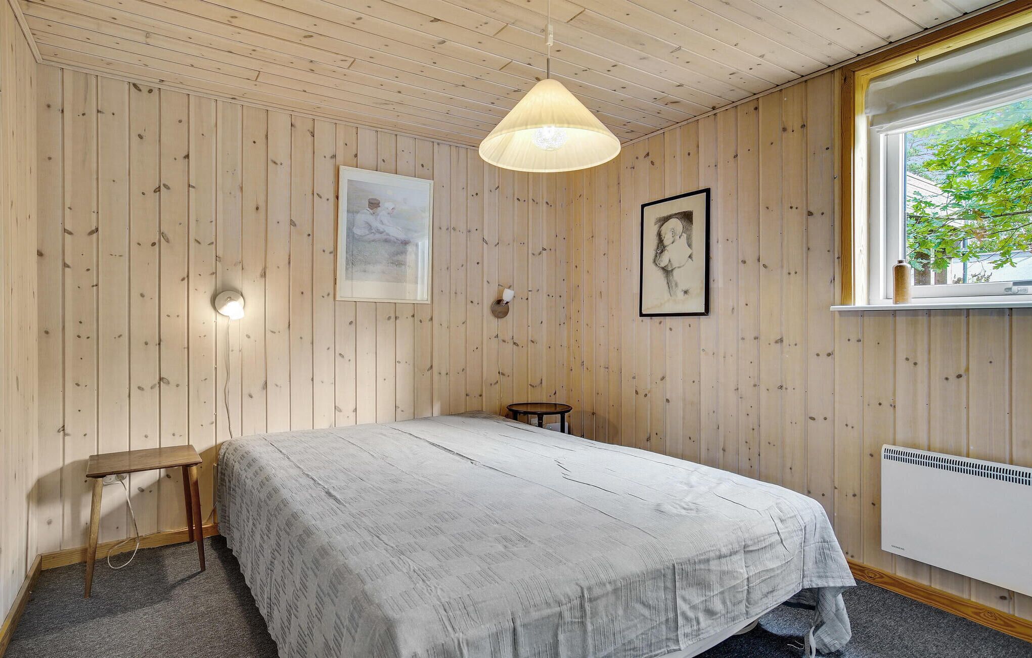 Ein Zimmer mit Holzwandverkleidung, einem Bett mit grauer Bettdecke, einer Hängelampe, einem kleinen Tisch und einem Fenster mit Blick auf Grünflächen.