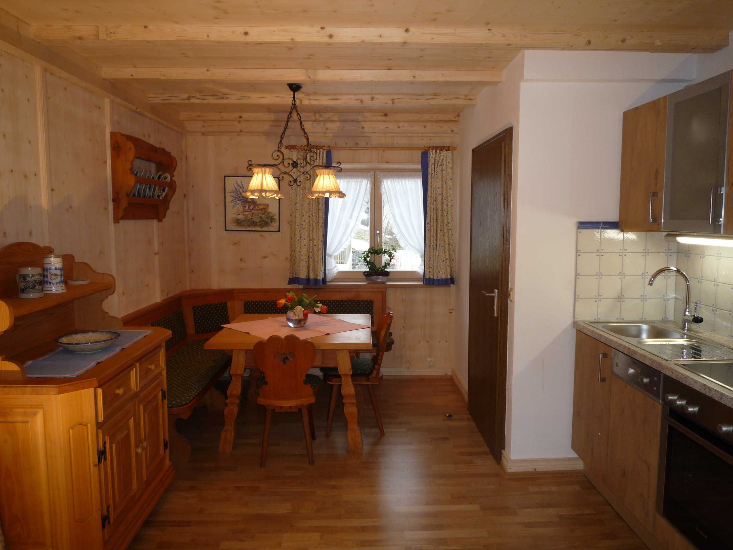 A wooden interior with a dining table, chairs, a chandelier, a kitchen with a sink and stove, and a window with curtains.