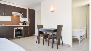 Une cuisine compacte avec des meubles sombres, un four encastré et une machine à café. On y trouve une table à manger avec deux chaises et un petit lit blanc dans un coin.